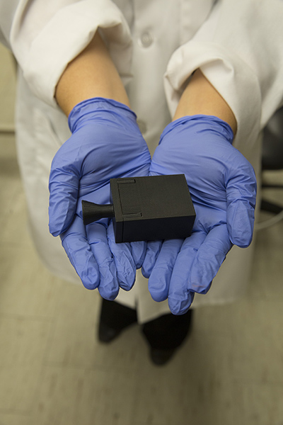 A photo of outstretched hands holding a small black device with a caption of "The device is a long way from its final form as an adhesive patch, but prototypes have gotten increasingly smaller since Associate Professor Jennifer Blain Christen first started the project. The initial version of the device was about the size of a shoebox and has progressed to a prototype mere inches across. Photographer: Pete Zrioka/ASU"