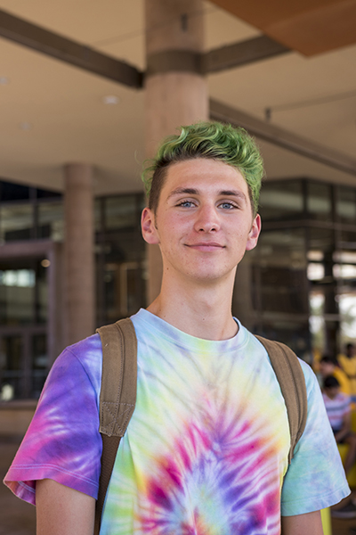 A young man in a tie-dyed shirt with green hair smiles for a photo. 