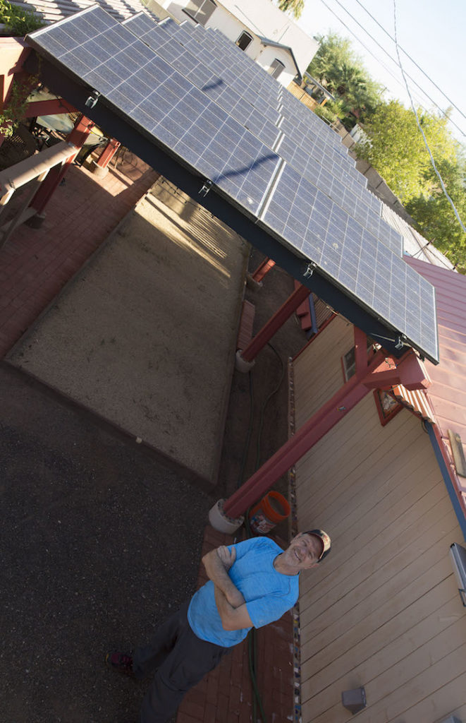 Photo of Ron Roedel standing below an array of solar panels. Caption: Ron Roedel stands near the “Power Pergola” in the back yard of his home in a historic neighborhood in Phoenix. The photovoltaic system atop a trellis structure supplies all of the electricity for his house. In recent years he has become more active in public advocacy to promote the development of solar power infrastructure as a renewable energy resource. Photography: Pete Zrioka/ASU