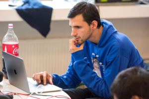 A young man with dark hair is hunched over a laptop, scratching his chin pensively. The caption reads: Software engineering graduate student Alexander Lampis Slaughter works on the computer vision challenge during the 2017 VisionHack, held in Moscow from September 11-13, 2017. The hackathon tasked teams with teaching a computer to recognize various obstacles and traffic occurrences for use in autonomous vehicles. Lampis Slaughter brought his experience with web development to the team. Photo courtesy of Jared Schoepf