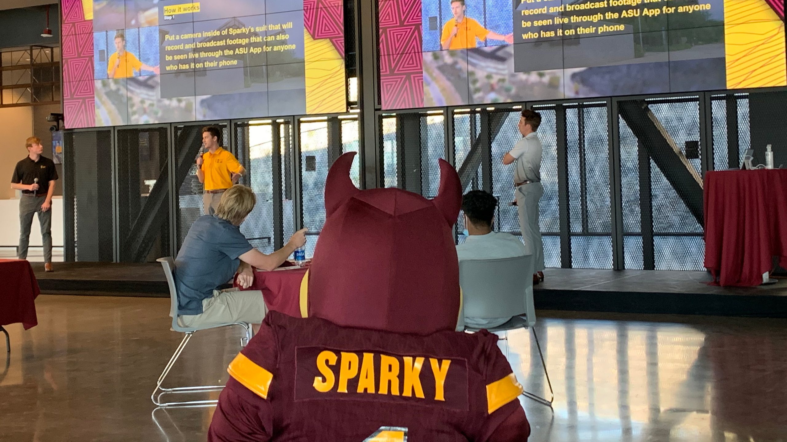 three students presenting a project on a stage during Ignite Sparky. With Sparky the Sun Devil sitting and watching.